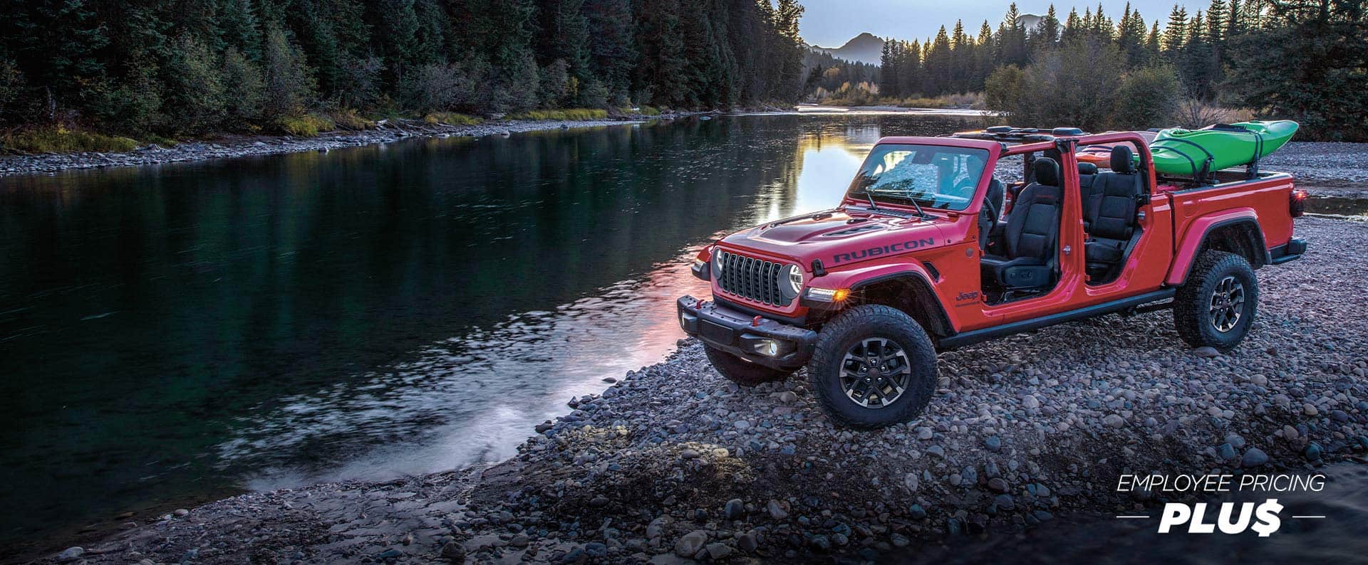 A red 2025 Jeep Gladiator Rubicon X with its doors and roof removed and a canoe in its pickup bed, parked on a rocky shore beside a mountain river. Employee Pricing Plus Event.