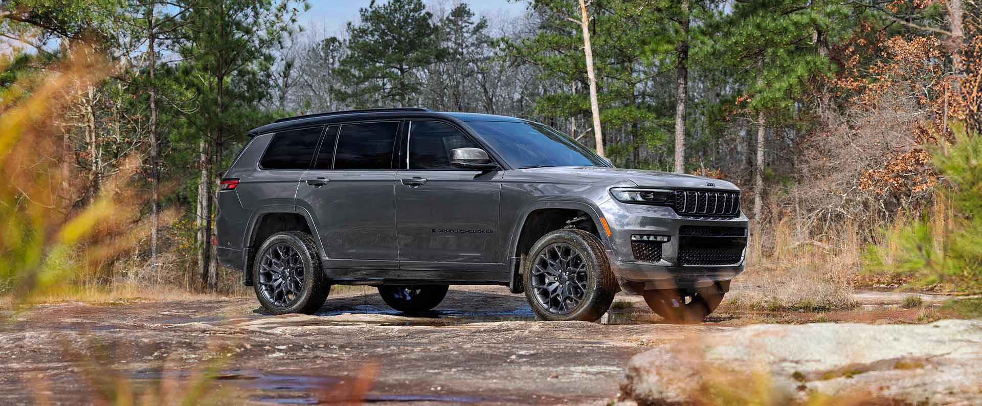 A gray 2025 Jeep Grand Cherokee L Summit Reserve High Altitude parked off-road beside a shallow, muddy stream.