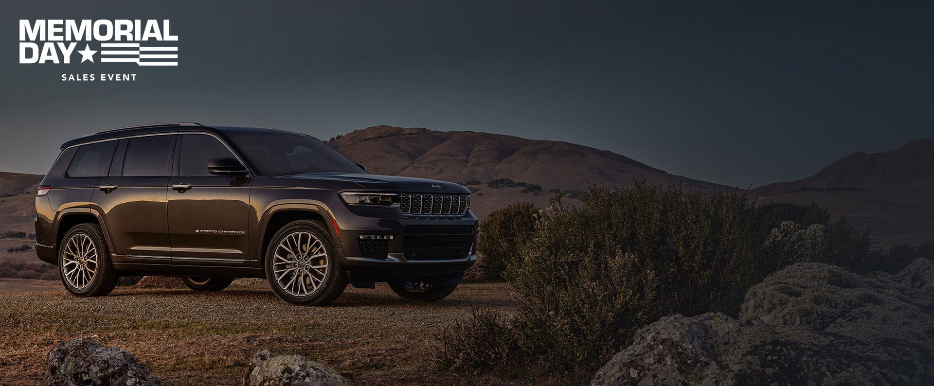 A passenger-side front angle of a gray 2025 Jeep Grand Cherokee Summit Reserve parked on a clearing off-road. Memorial day.