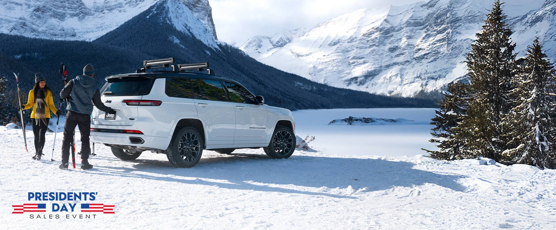 Un Jeep Grand Cherokee L High Altitude 2026 blanco estacionado en un claro cubierto de nieve en las montañas. Una pareja cerca del vehículo está transportando equipo de esquí, y el hombre está a punto de abrir la puerta levadiza. El evento de ventas por el Día del Presidente.