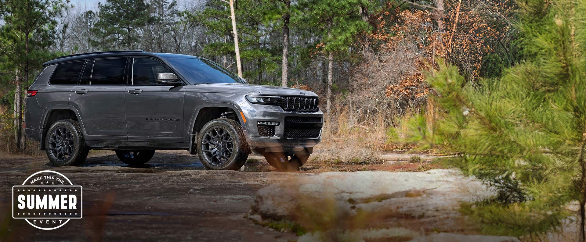 A gray 2025 Jeep Grand Cherokee L Summit Reserve High Altitude parked off-road beside a shallow, muddy stream.