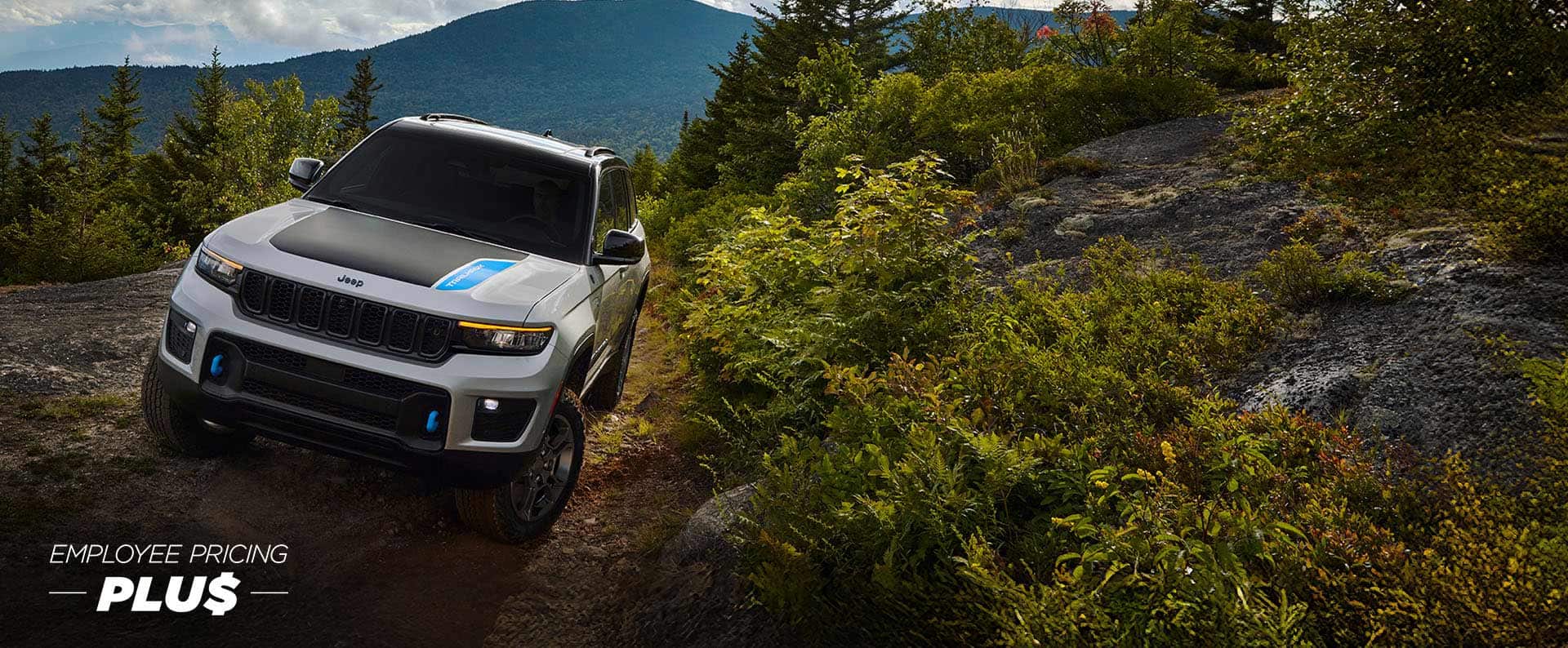 A white 2025 Jeep Grand Cherokee 4xe Trailhawk with black hood insert, crawling on a trail off-road in the mountains. Employee pricing plus event.