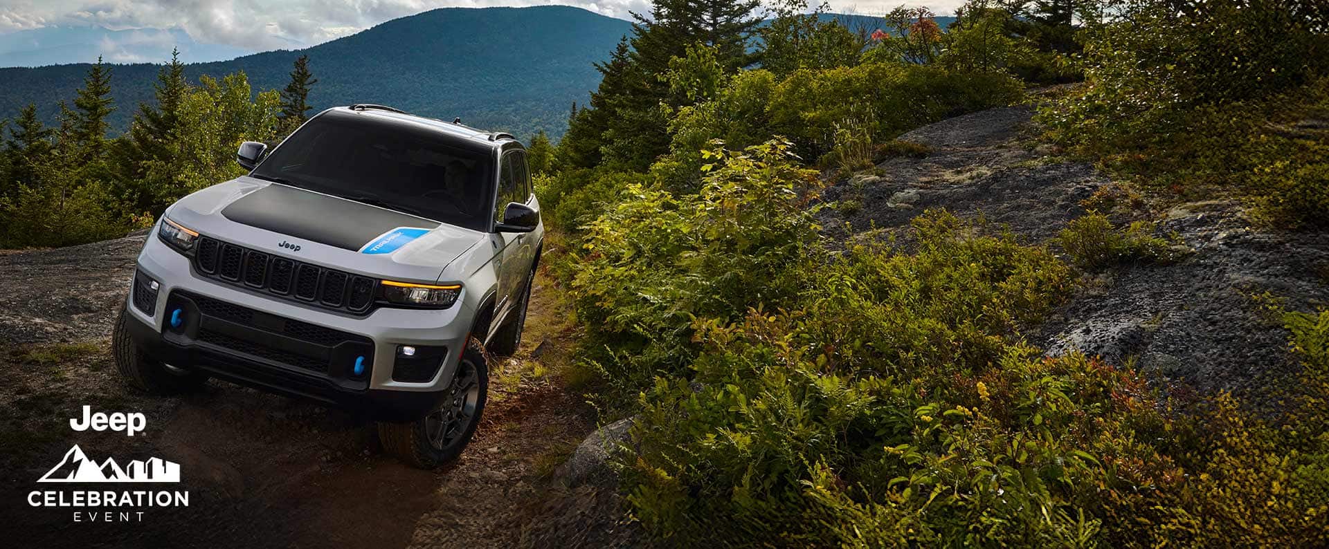A white 2025 Jeep Grand Cherokee 4xe Trailhawk with black hood insert, crawling on a trail off-road in the mountains. Jeep Celebration Event.