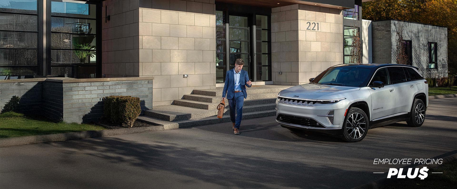 A silver 2025 Wagoneer S Limited parked in the driveway of a commercial building with a man walking nearby. Employee Pricing Plus event.