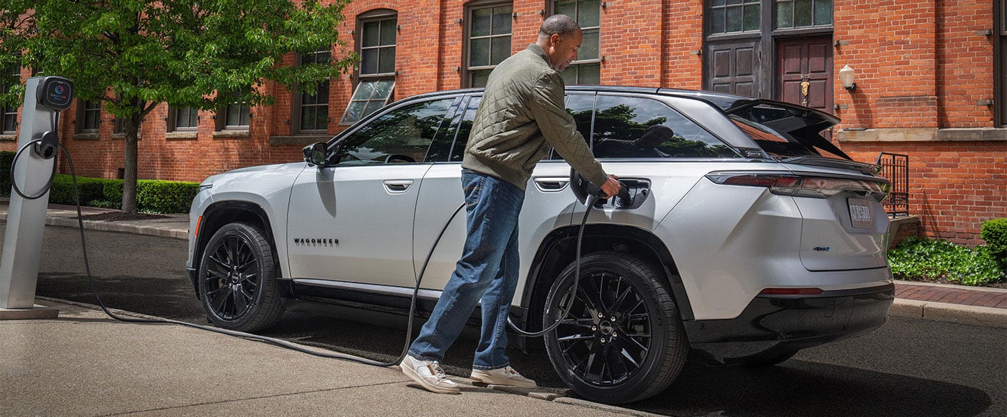 A silver 2025 Jeep Wagoneer S Launch Edition with a man plugging  the charging cord from a public charging station into the vehicle's charging port.