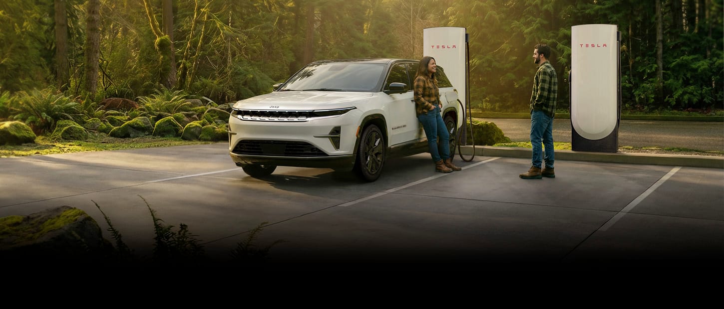 Un Jeep Wagoneer S Launch Edition 2026 blanco estacionado en una estación de carga Tesla en un bosque denso, con una pareja de pie fuera del vehículo y un cable de carga del Supercargador enchufado al puerto de carga del Wagoneer S.