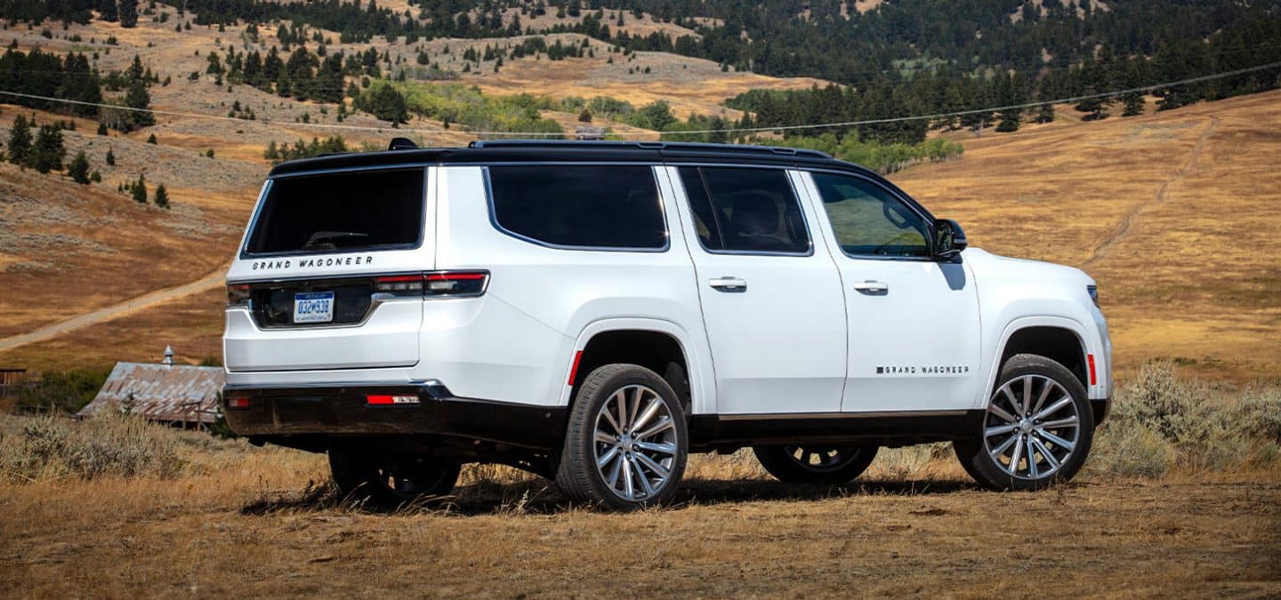 Display A passenger-side rear angle of a white 2025 Jeep Grand Wagoneer Series 3 parked in an open field in an area of hilly grasslands.