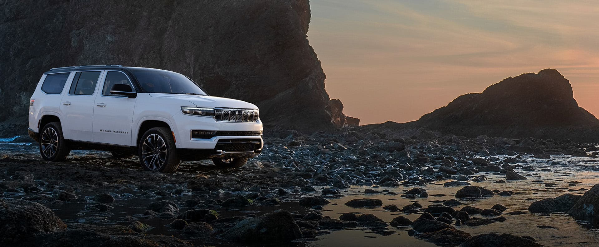 A passenger-side front angle of a white 2025 Jeep Grand Wagoneer Series 3 parked on a rocky beach. Drive Into Fall Event.