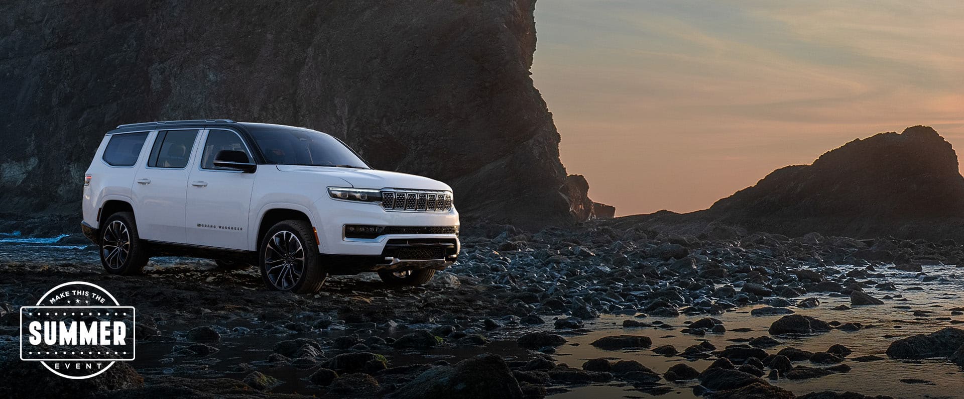 A passenger-side front angle of a white 2025 Jeep Grand Wagoneer Series 3 parked on a rocky beach. Make This The Summer Event.