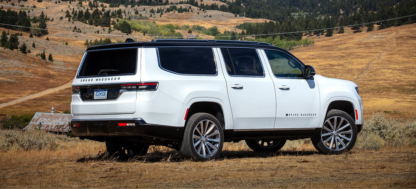 A passenger-side rear angle of a white 2025 Jeep Grand Wagoneer Series 3 parked in an open field in an area of hilly grasslands.