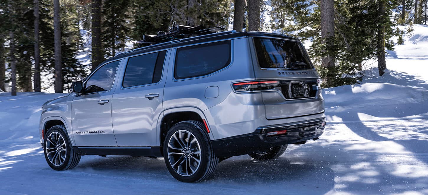 A driver-side rear angle of a white 2025 Jeep Grand Wagoneer Series 3 traveling on a snow-covered trail in the woods, off-road.
