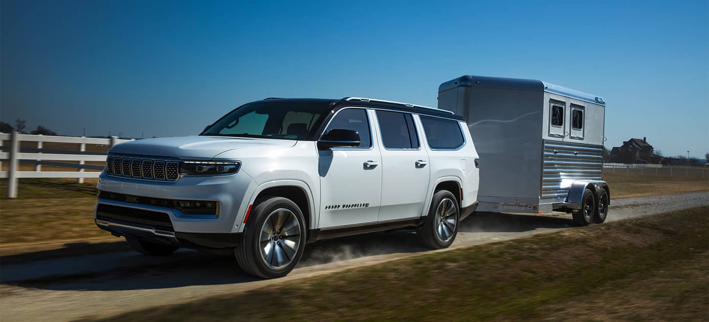 A white 2025 Jeep Grand Wagoneer Series 3 traveling down a dirt road beside a farm, towing a horse trailer.