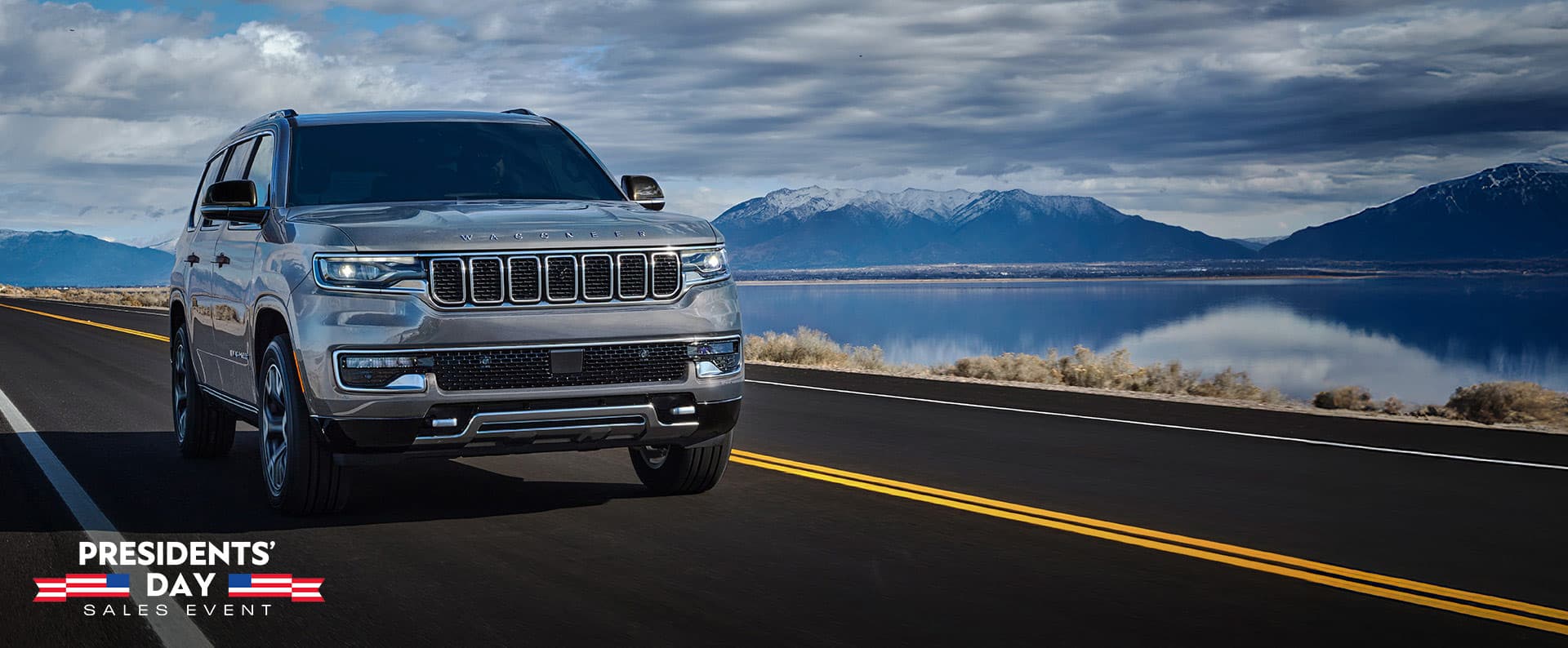 Un Jeep Wagoneer Series 3 2025 plateado circulando por una carretera al lado de un lago. El evento de ventas por el Día del Presidente.