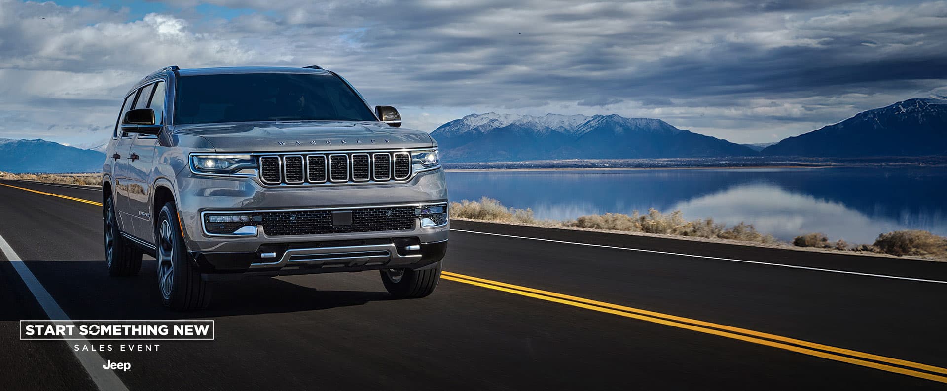 Un Jeep Wagoneer Series 3 2025 plateado circulando por una carretera al lado de un lago, en invierno. El evento de ventas Comienza algo nuevo. Jeep