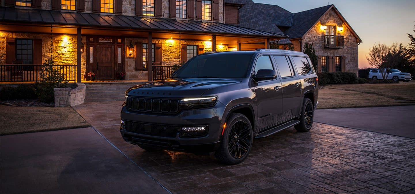 Display A driver-side front angle of a black 2025 Jeep Wagoneer Carbide with its Daytime Running Lamps on, parked in the courtyard of a large, upscale lodge at sunset.