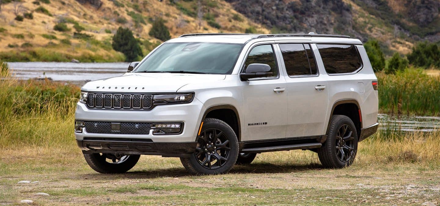 Display A driver-side front angle of a white 2025 Jeep Wagoneer Carbide parked on a grassy clearing beside a swampy river.