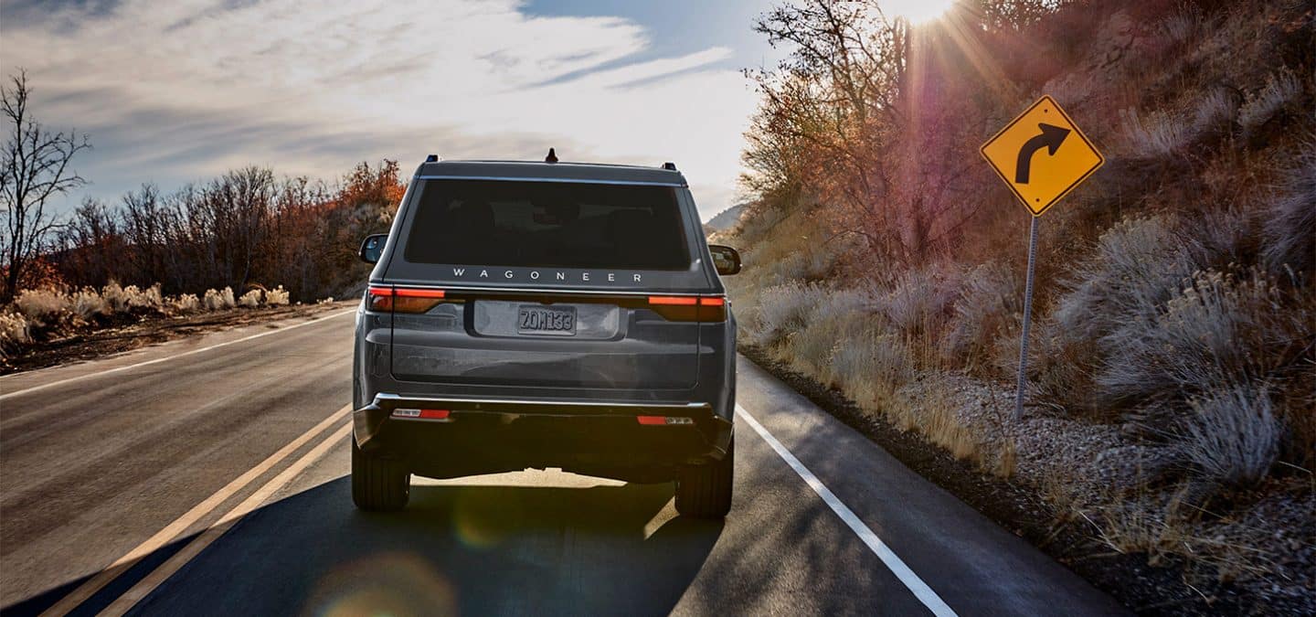 Display A rear angle of a gray 2025 Jeep Wagoneer Series III traveling on a curved highway in the mountains.