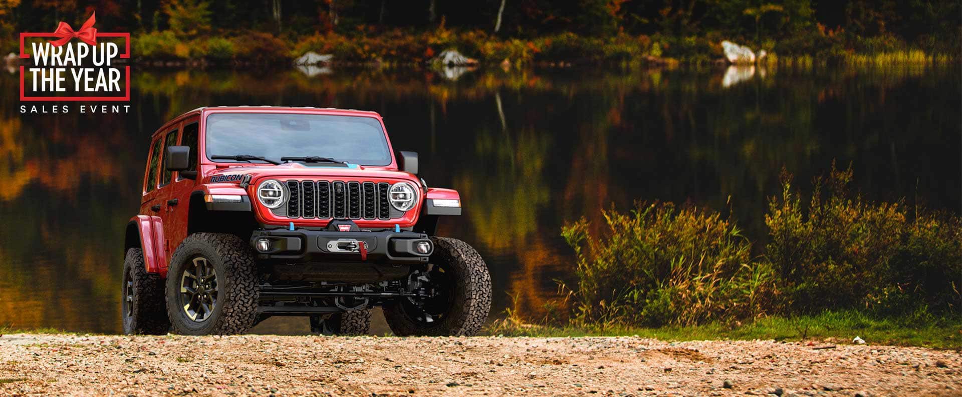 A red 2025 Jeep Wrangler Rubicon X 4xe parked on a hill off-road, with a lake in the background. The Drive Into Fall Event.