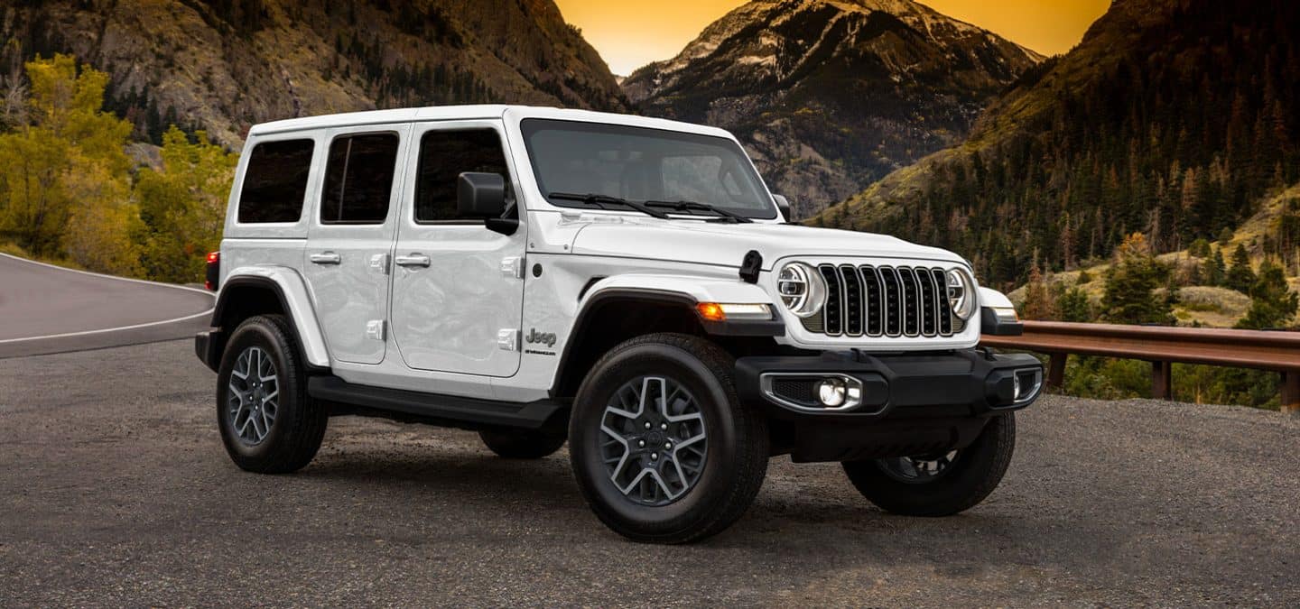 Display A passenger-side front angle of a white 2025 Jeep Wrangler Sahara parked on the shoulder of a highway in the mountains at sunset.