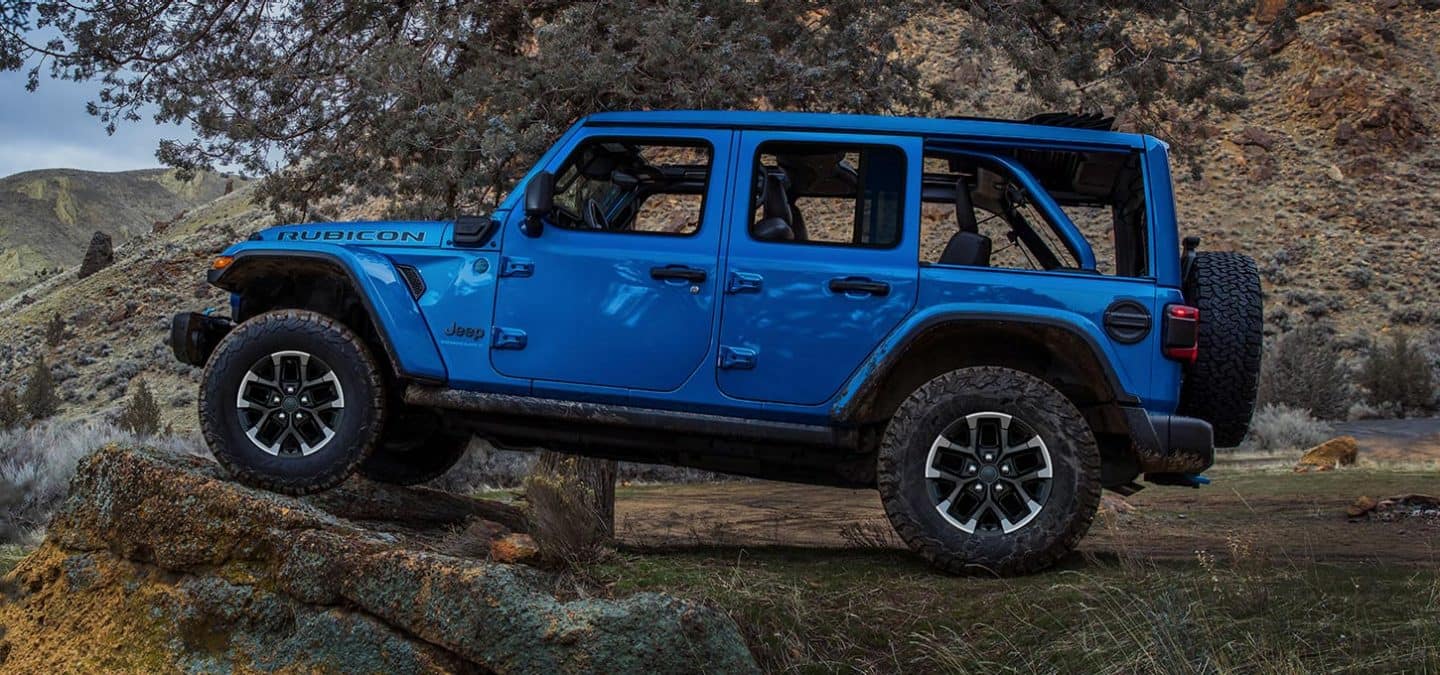 Display A driver-side profile of a blue 2025 Jeep Wrangler Rubicon X 4xe parked off-road, with its front tires elevated on a raised rock formation.