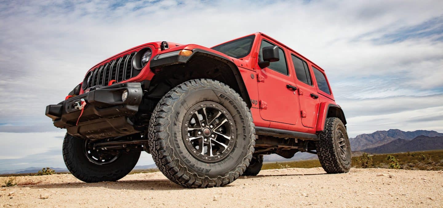 Display A low front angle of a red 2025 Jeep Wrangler Rubicon X parked on sand, with mountains in the background.