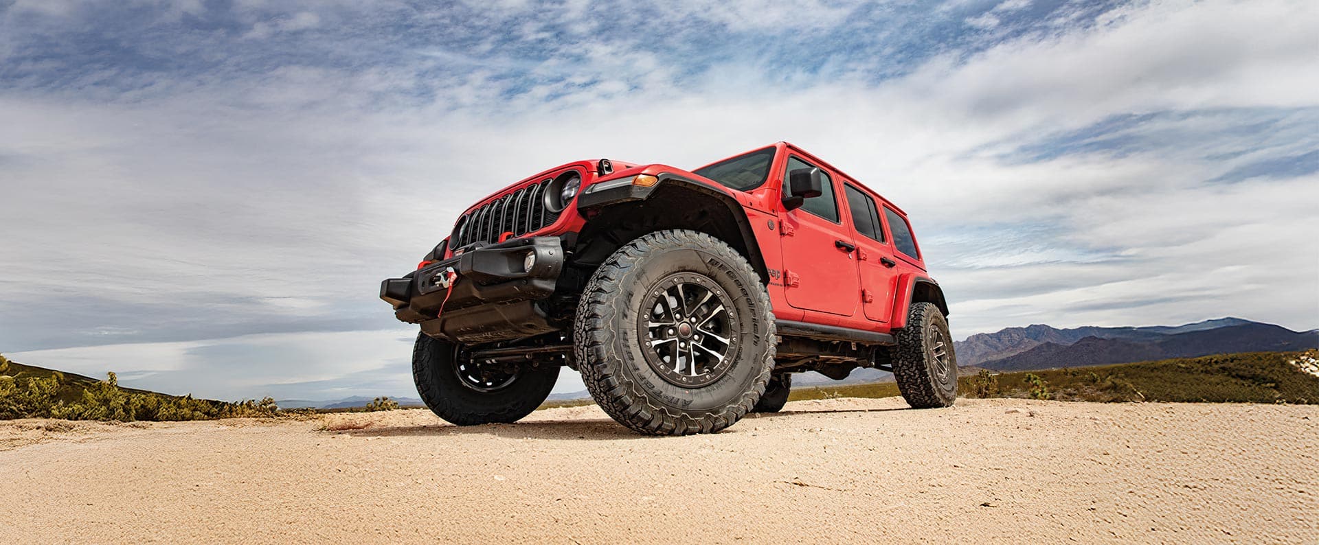 A low-slung front angle of a red 2025 Jeep Wrangler Rubicon X traveling on a sandy trail off-road.