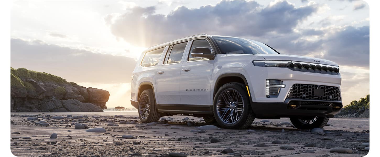 A lowered passenger-side front angle of a white 2026 Jeep Grand Wagoneer 85th Anniversary Edition, parked on a rocky beach at sunset.