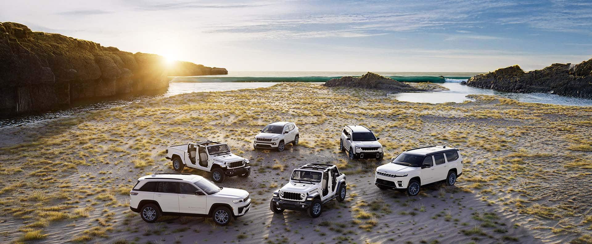 Six 2026 Jeep 85th Anniversary Edition vehicles, all in white, parked in a semicircle on a beach at sunset, with Wrangler in the middle of the circle. Beginning on the left: Grand Cherokee, Gladiator, Compass, Cherokee and Grand Wagoneer. Both Wrangler and Gladiator have their doors and tops removed.
