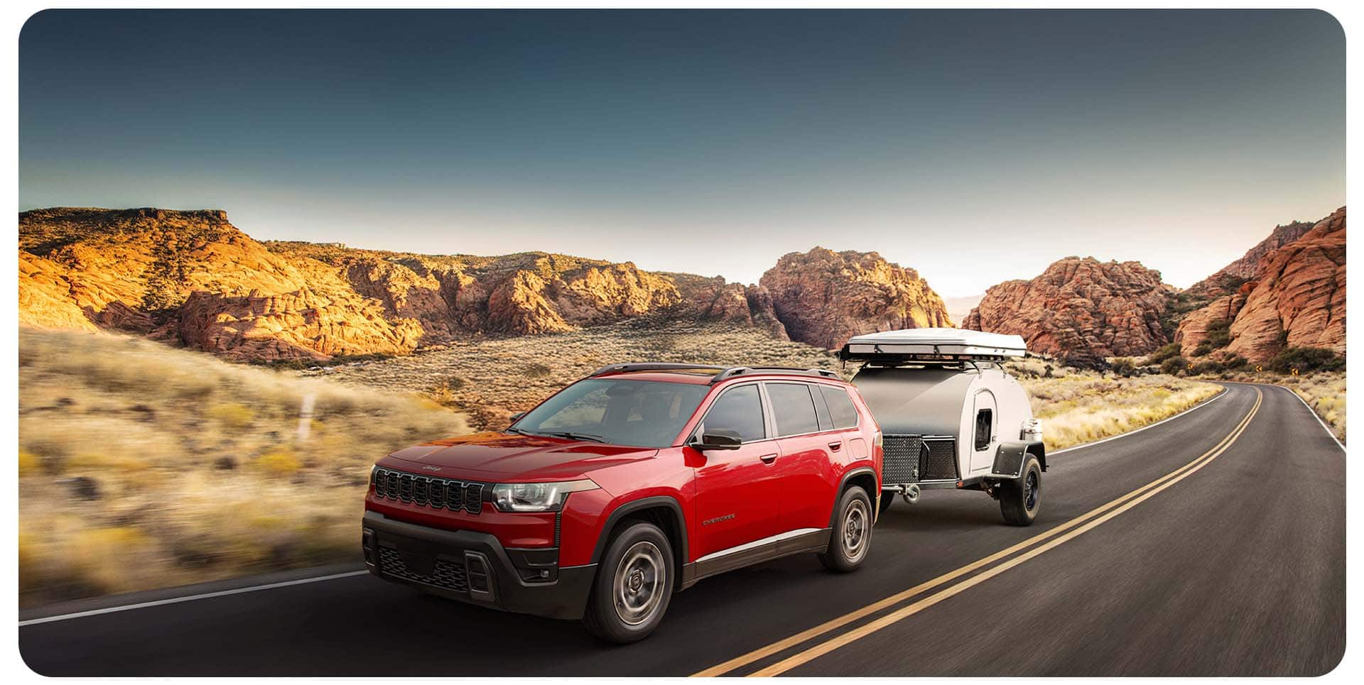 Un Jeep Cherokee Limited 2026 rojo circulando por una carretera, acarreando un pequeño remolque de viaje.