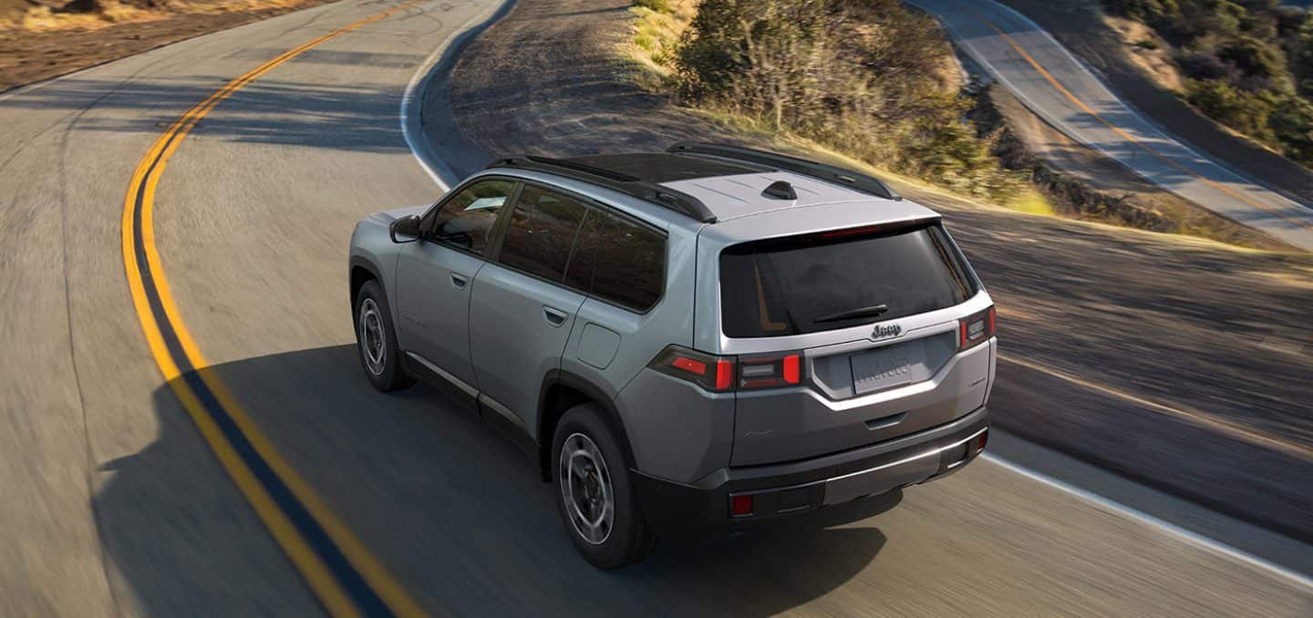 Display A driver-side rear angle of a silver 2026 Jeep Cherokee Limited traveling down a winding highway.