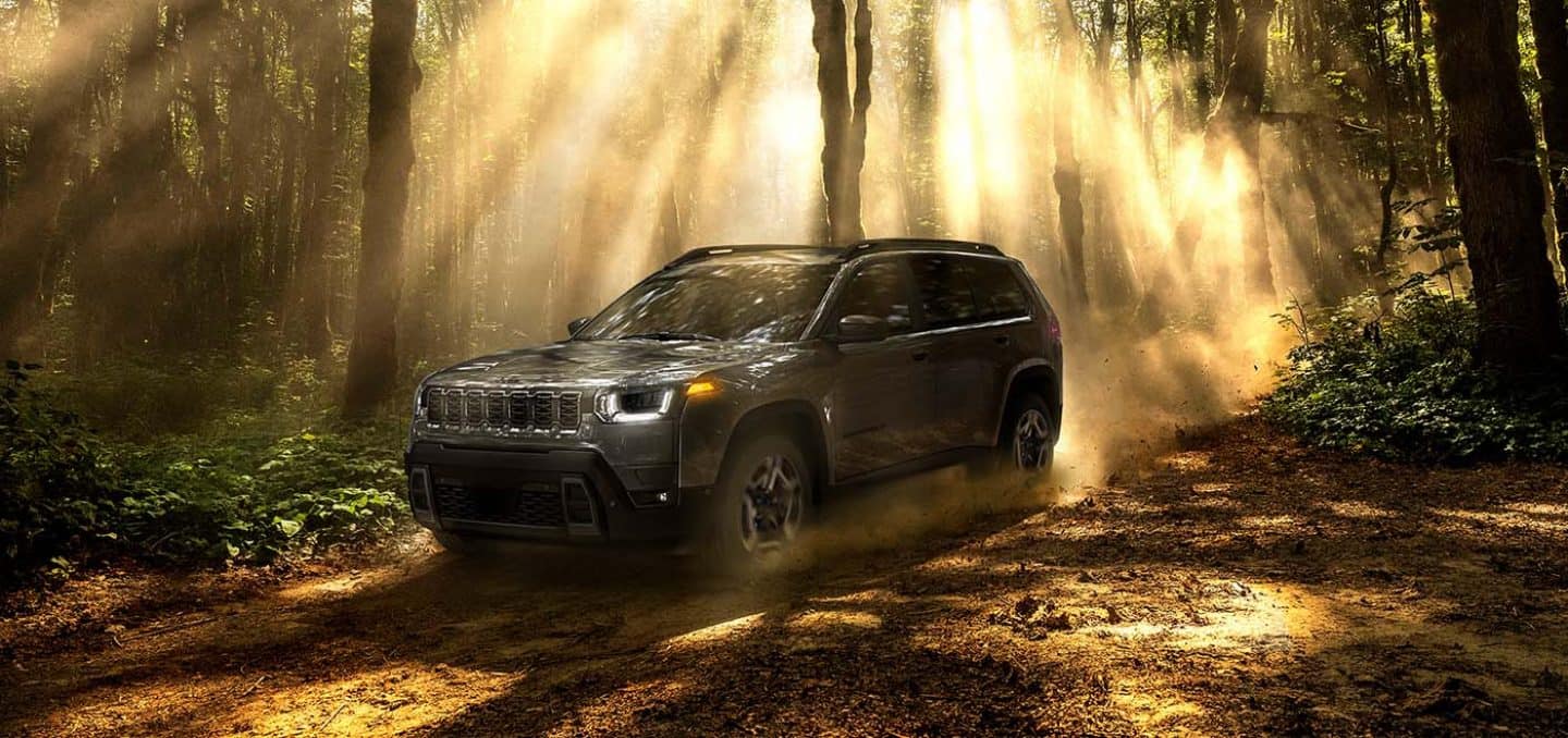 Display A driver-side front angle of a gray 2026 Jeep Cherokee Limited traveling on a trail off-road, through a forest with sunbeams shining through the trees.