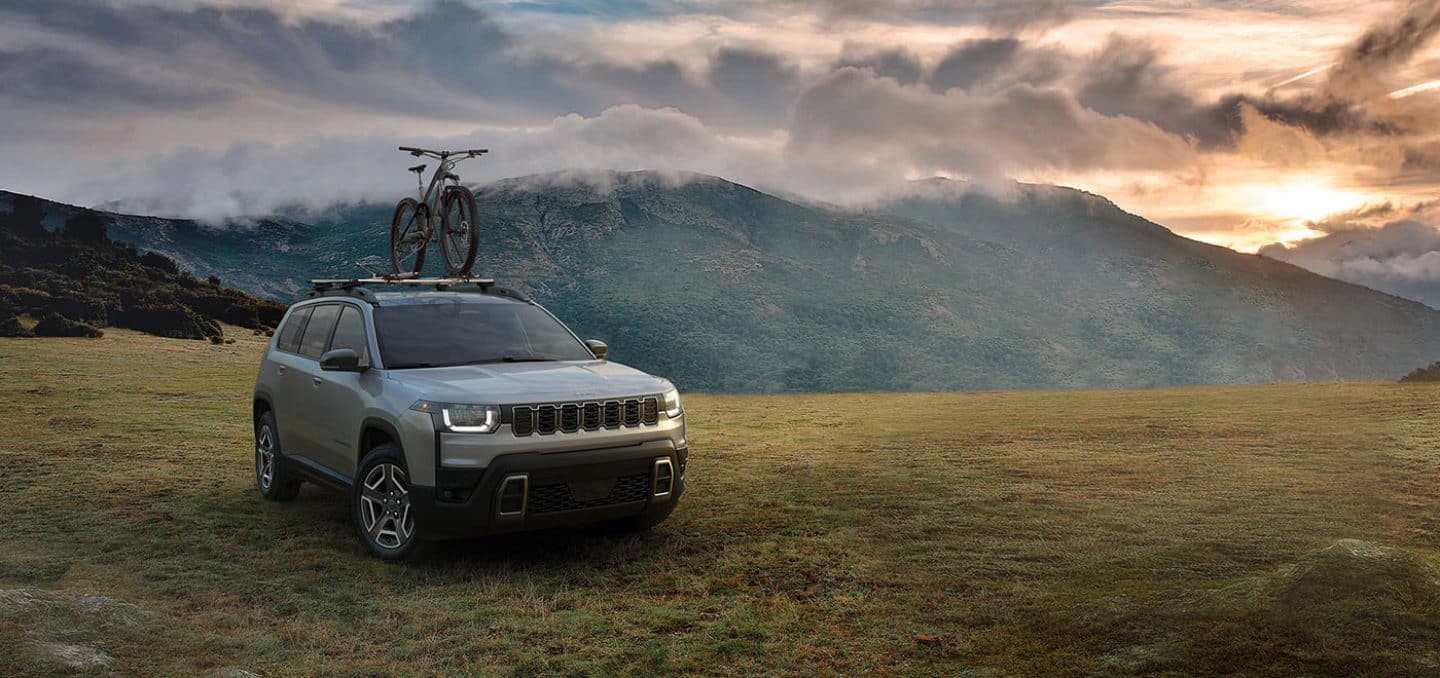Display A silver 2026 Jeep Cherokee Laredo with a bike strapped to its roof rack, parked in a field with mountains and dark clouds in the background.