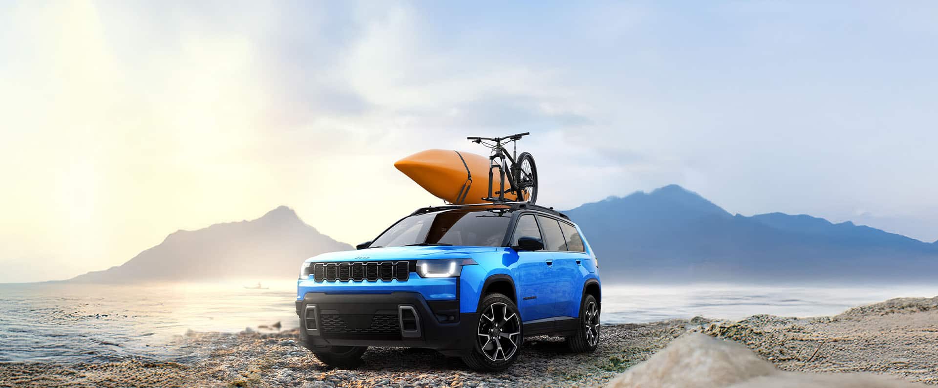 A blue 2026 Jeep Cherokee Limited parked on a rocky beach with a kayak and part of a bike strapped to its roof rack.