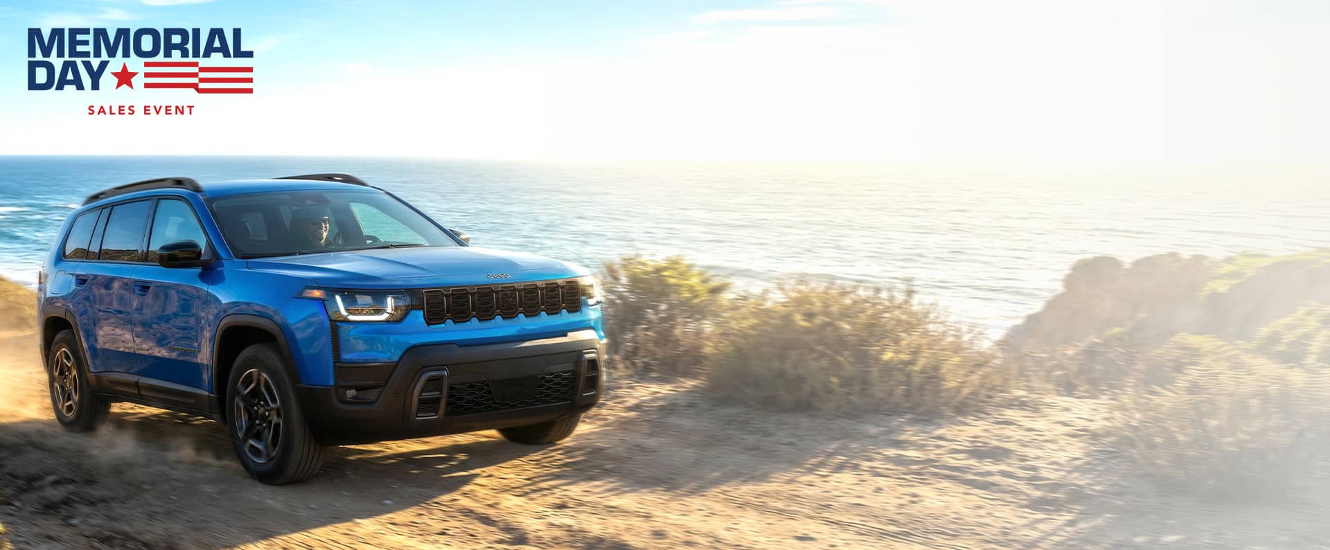 A passenger-side front angle of a blue 2026 Jeep Cherokee Laredo traveling on a beach beside the ocean. Memorial Day Sales Event.