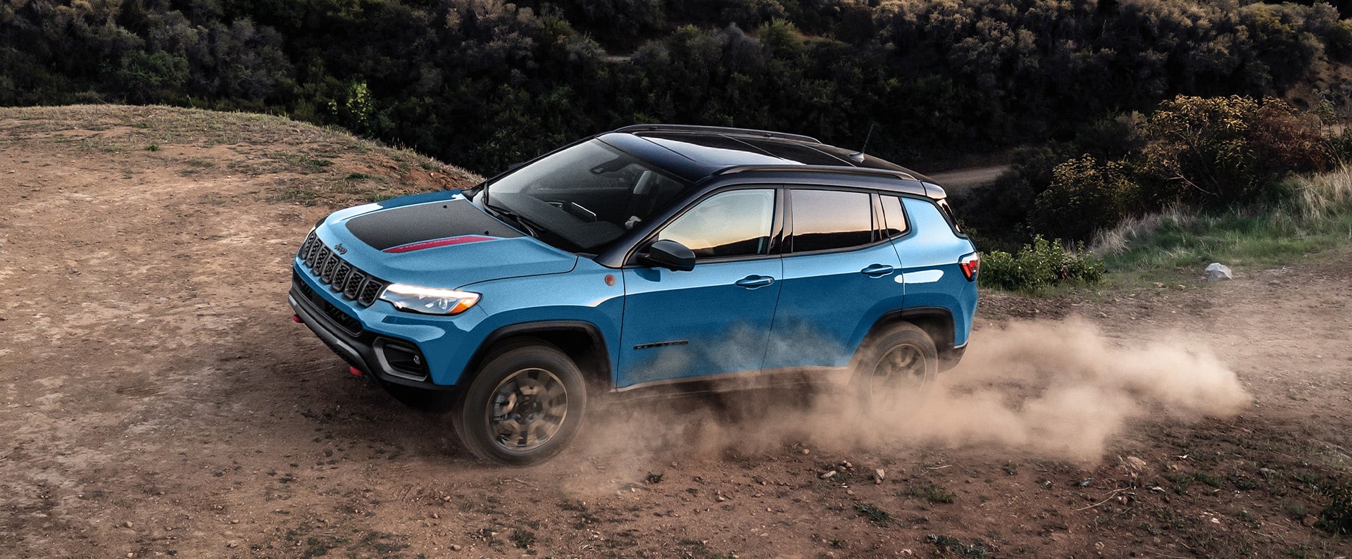 A blue 2026 Jeep Compass Trailhawk with a black hood insert and black roof, ascending a hill on a trail off-road, with the tires kicking up dust.