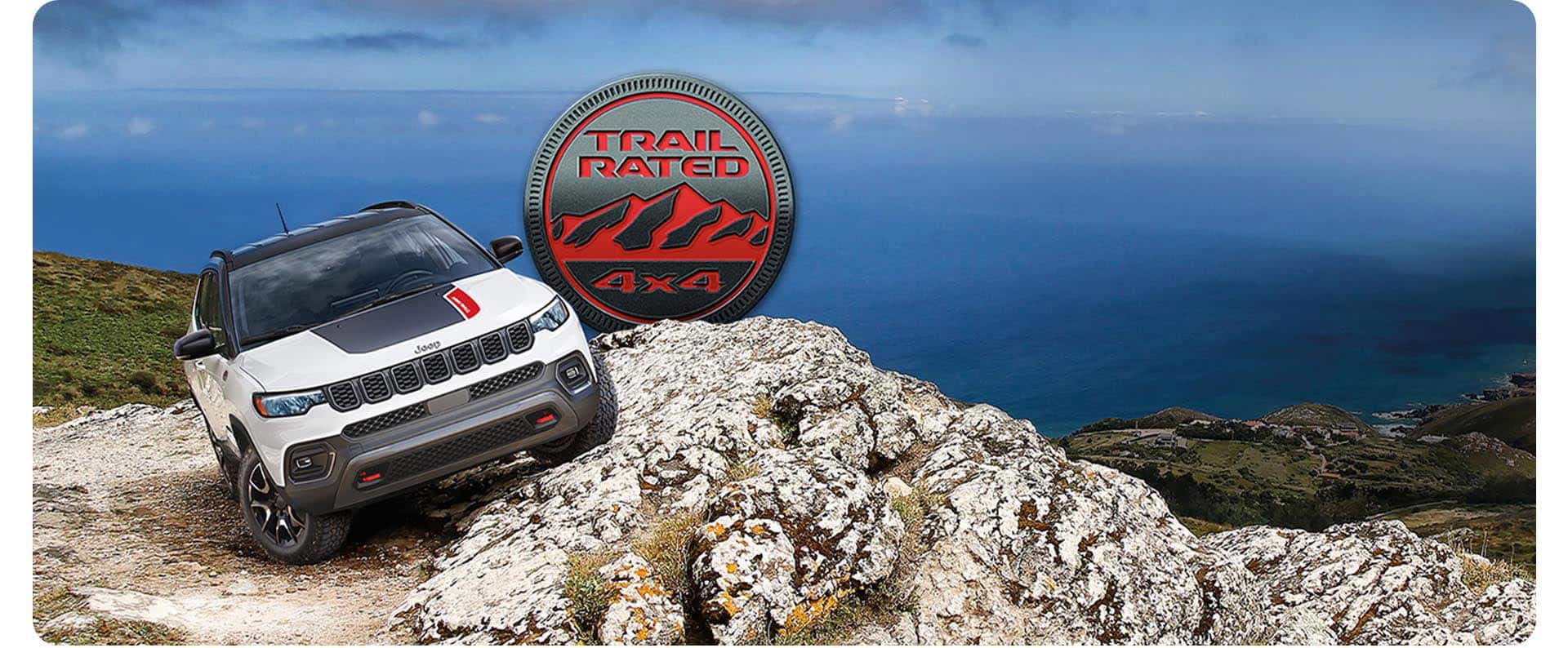 A white 2026 Jeep Compass Trailhawk with a black hood insert, parked horizontally on a steep, rocky hill, with the driver-side of the vehicle elevated on the uneven surface. Trail Rated 4x4 badge.