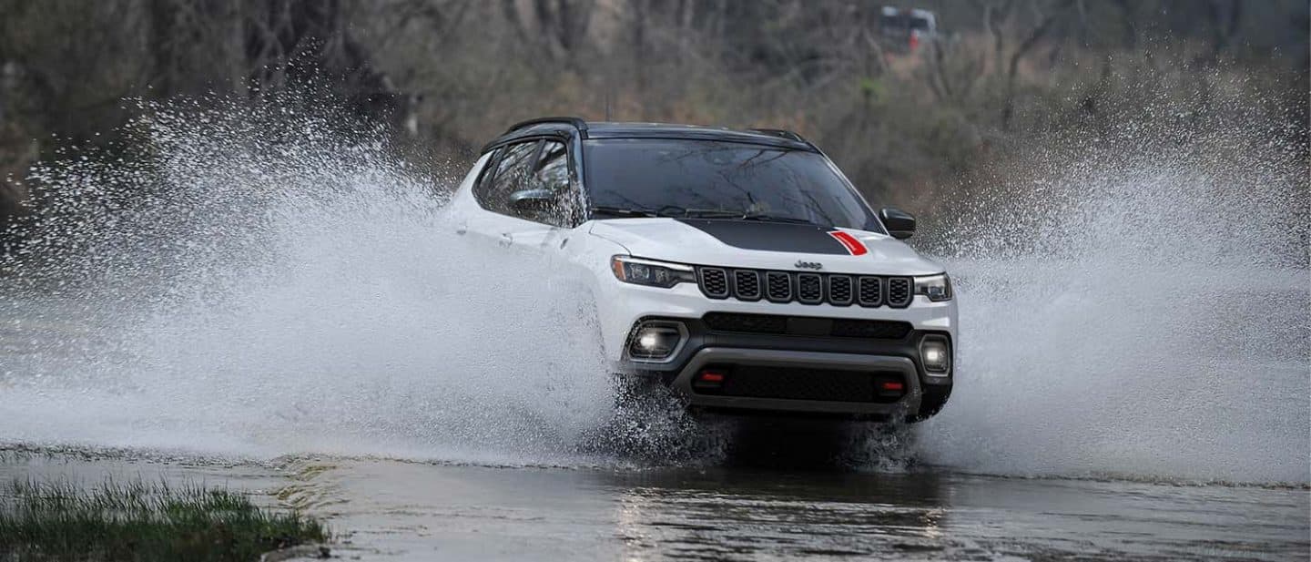 Display A white 2026 Jeep Compass Trailhawk with a black hood insert and black roof, fording through a stream with water splashing up the sides of the doors.