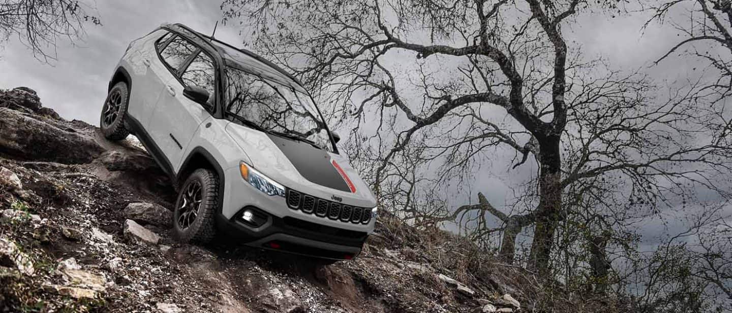 Display A white 2026 Jeep Compass Trailhawk with a black hood insert and black roof, descending an extremely steep hill off-road.