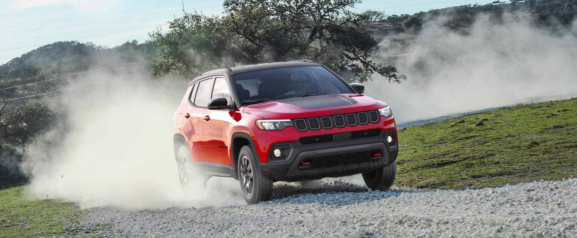 A red 2026 Jeep Compass Trailhawk traveling up a rocky trail off-road, kicking up dust as it goes.