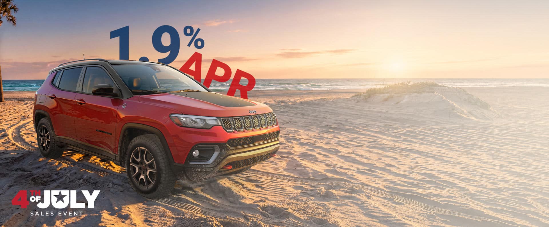 A red 2026 Jeep Compass Trailhawk with black hood insert, parked on a sandy beach at sunset. 1.9% APR. The 4th of July Sales Event.