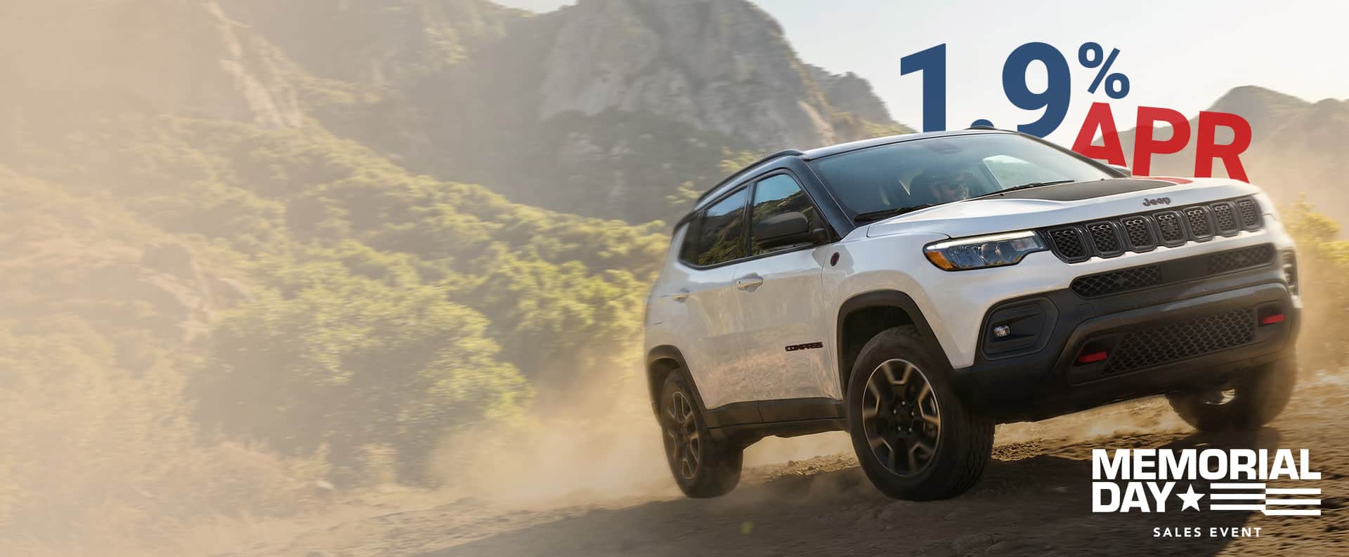 A passenger-side front angle of a white 2026 Jeep Compass Trailhawk with black hood insert ascending a trail off-road. Memorial Day Sales Event.