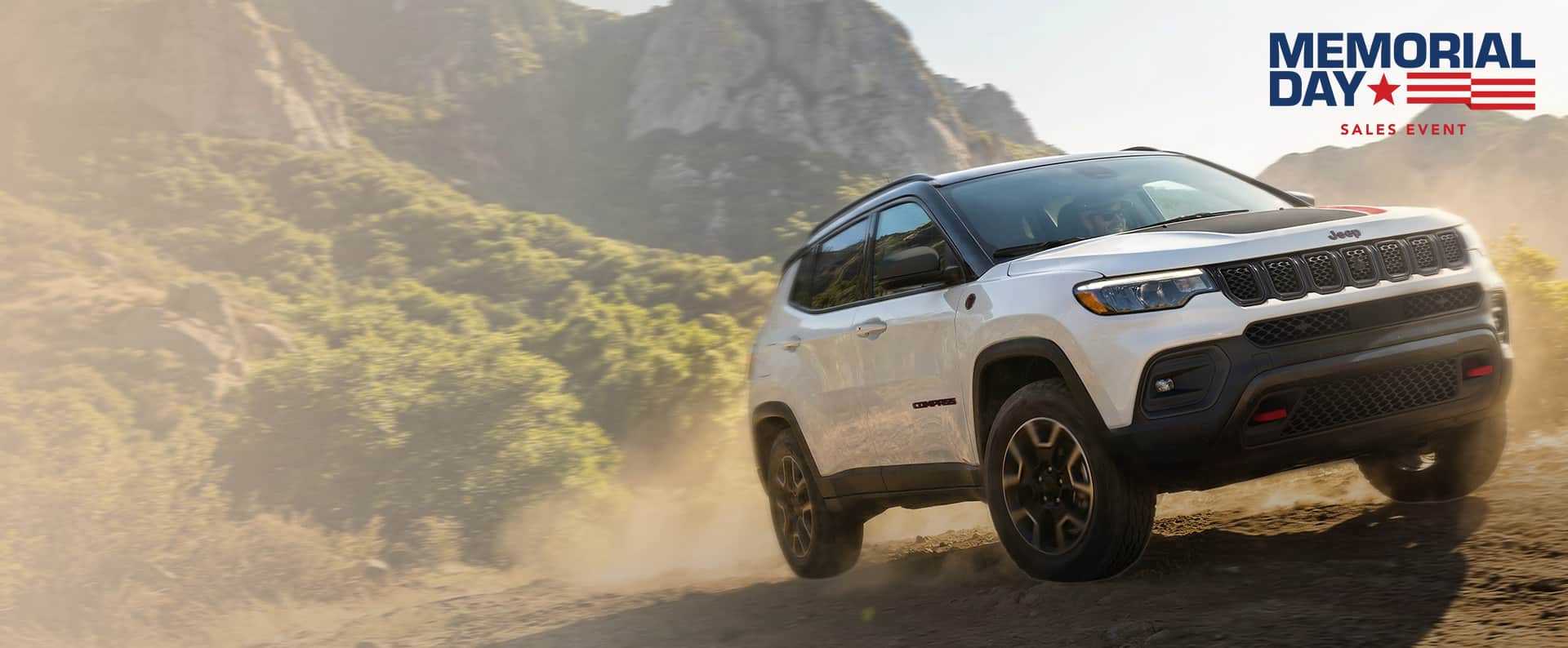 A passenger-side front angle of a white 2026 Jeep Compass Trailhawk with black hood insert ascending a trail off-road. Memorial Day Sales Event.