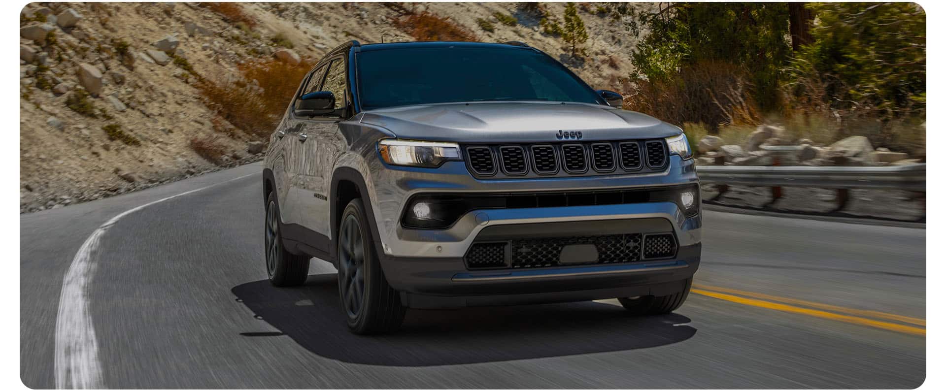 A front angle of a silver 2026 Jeep Compass Limited Altitude traveling on a winding mountain highway.