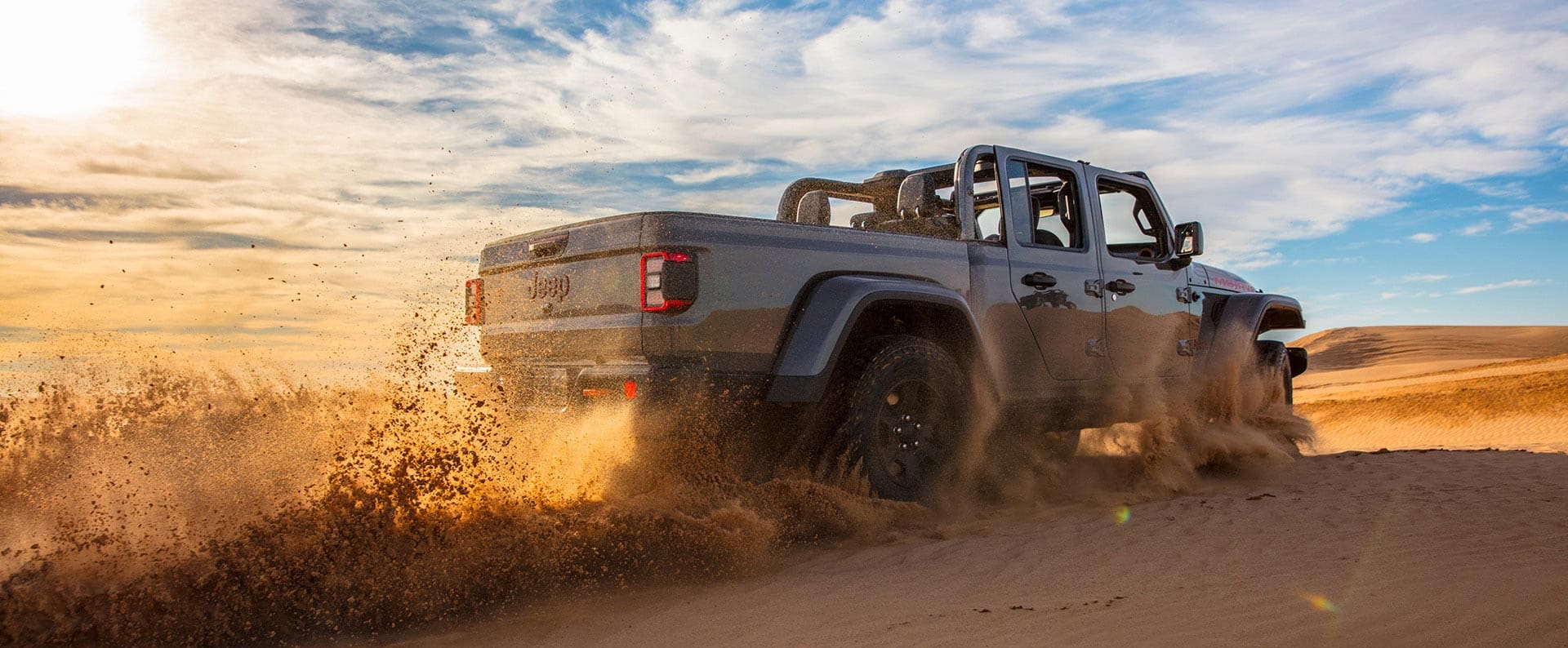 A passenger-side rear angle of a gray 2026 Jeep Gladiator Mojave X with its doors and roof removed, traveling through the desert at sunset, kicking up sand as it goes.