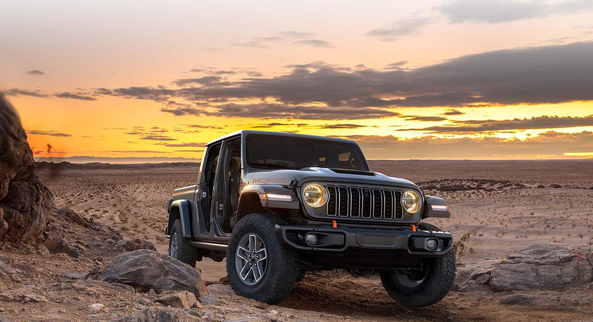 A gray 2026 Jeep Gladiator Mojave with its doors removed, ascending a hill in the desert at sunset.