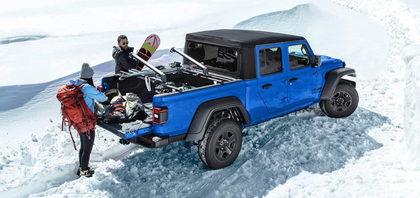 Display A passenger-side rear angle of a blue 2026 Jeep Gladiator Sport with a black top and its tailgate open, parked on the snow with a couple pulling winter sports equipment out of the pickup box.