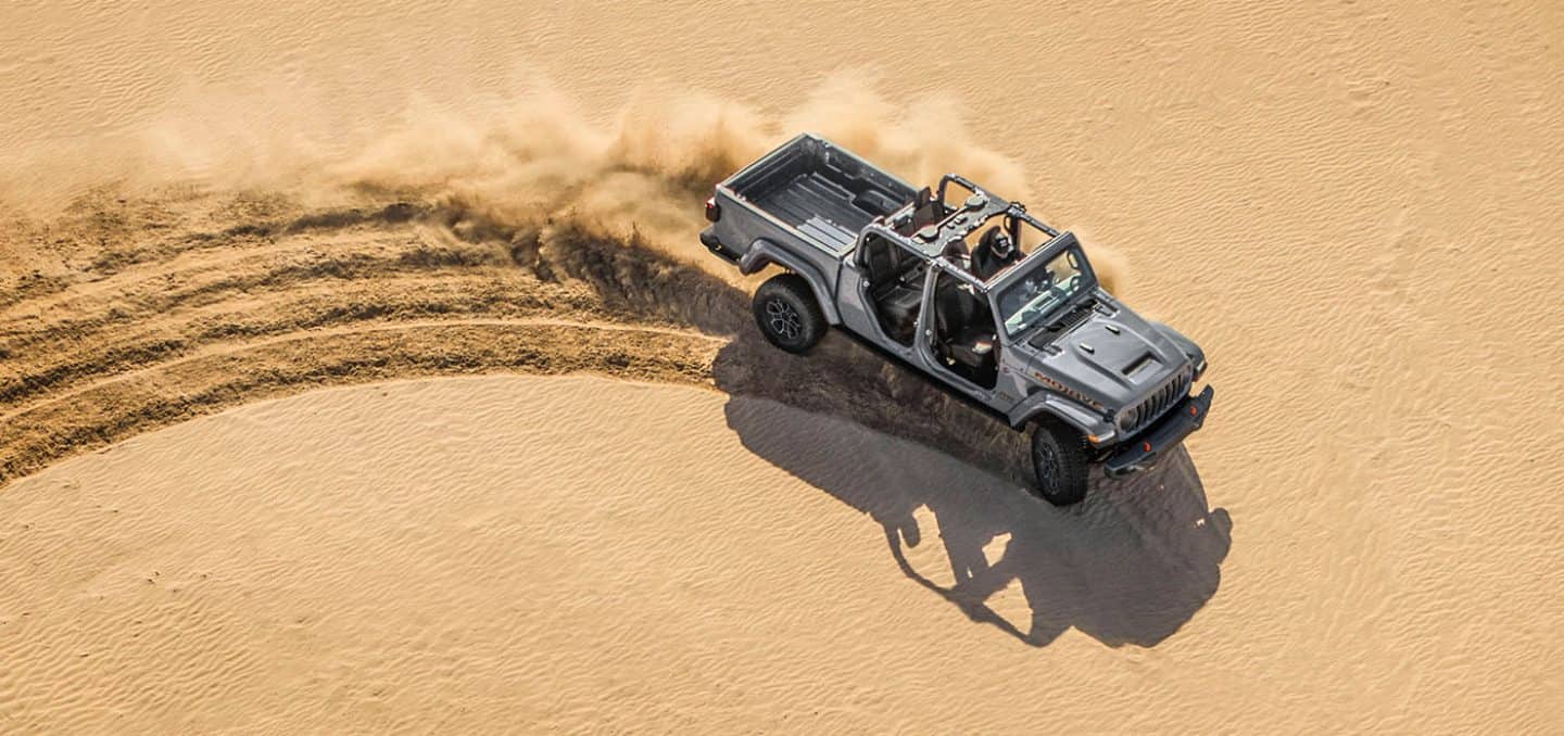 Display A overhead view of a gray 2026 Jeep Gladiator Mojave X with its doors and top removed, being driven in loose sand, creating heavy tracks as it goes.