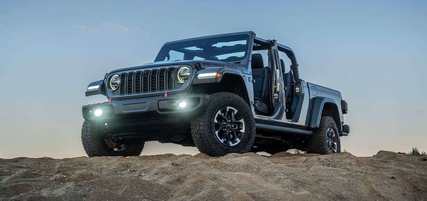 Display A gray 2026 Jeep Gladiator Rubicon X with its doors and top removed and its headlamps and fog lamps on, parked on the top of a rocky hill.