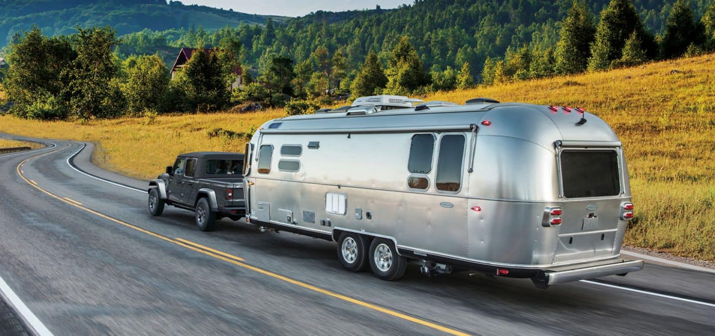 Display A gray 2026 Jeep Gladiator Sport S traveling down a highway, towing a large travel trailer, with mountains in the background.