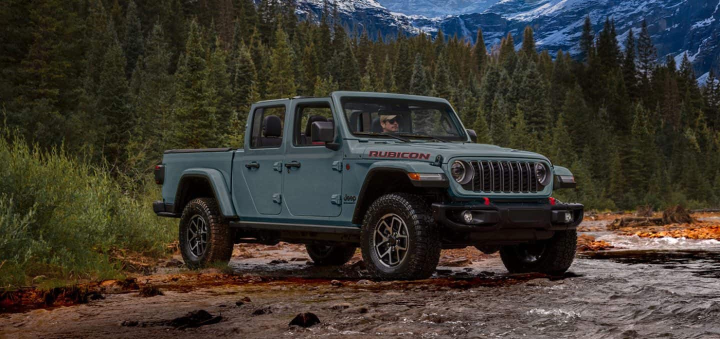Display A passenger-side front angle of a gray 2026 Jeep Gladiator Rubicon X entering a stream in a mountain forest.